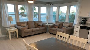 Sala de estar con dos sofás, una mesa de comedor y sillas. Cuenta con ventanas grandes con cortinas, una lámpara en una mesita auxiliar y una cafetera sobre la encimera.