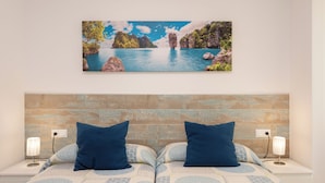 A double bed with a patterned bedspread, two blue pillows, and a nightstand with a lamp. A framed picture of a coastal landscape hangs above the bed. - Apartment with sea view, parking, wifi (Marbella)