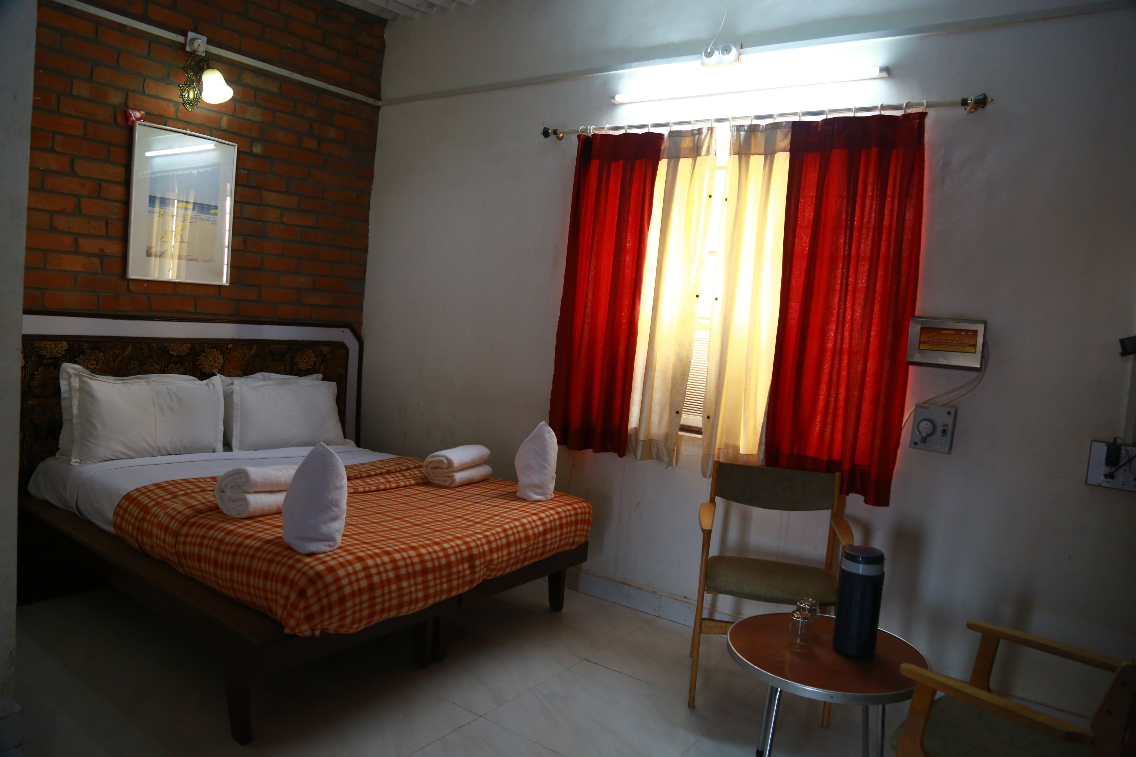 A hotel room with a bed, a chair, a table, a window with curtains, and a wall-mounted light fixture.