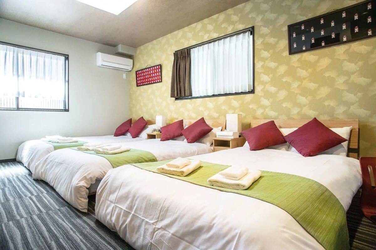 A hotel room with two beds, each with white linens, green bedspreads, and red pillows. There is a wall-mounted air conditioner, a window with white curtains, and a wall decoration above the beds.