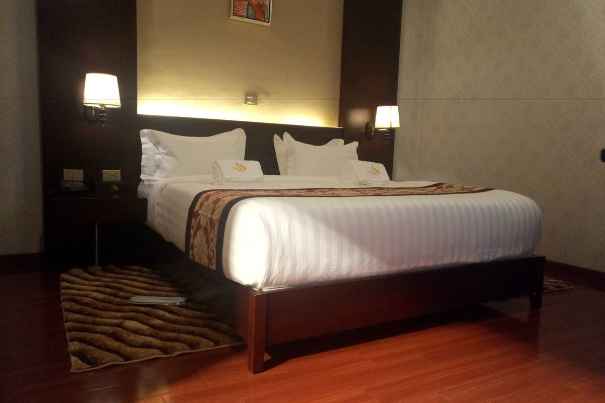 A hotel room with a large bed, two bedside tables, a nightstand, a wall-mounted lamp, and a patterned rug.