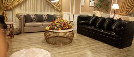 A living room with a beige sofa, a black leather sofa, a round coffee table with a floral centerpiece, and a lamp.