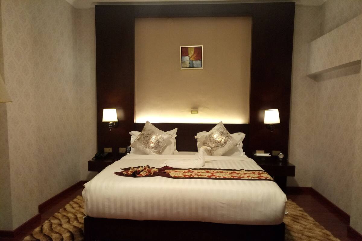 A hotel room with a large bed, two bedside lamps, a headboard with a decorative panel, and a small painting above the headboard.