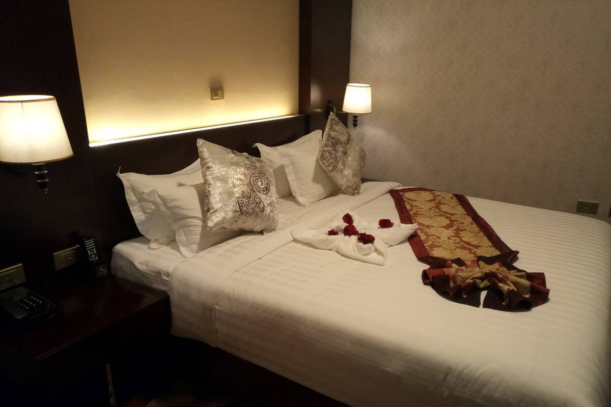 A neatly made bed with white linens, decorative pillows, and a red and gold throw blanket. There are two bedside lamps and a remote control on the nightstand.