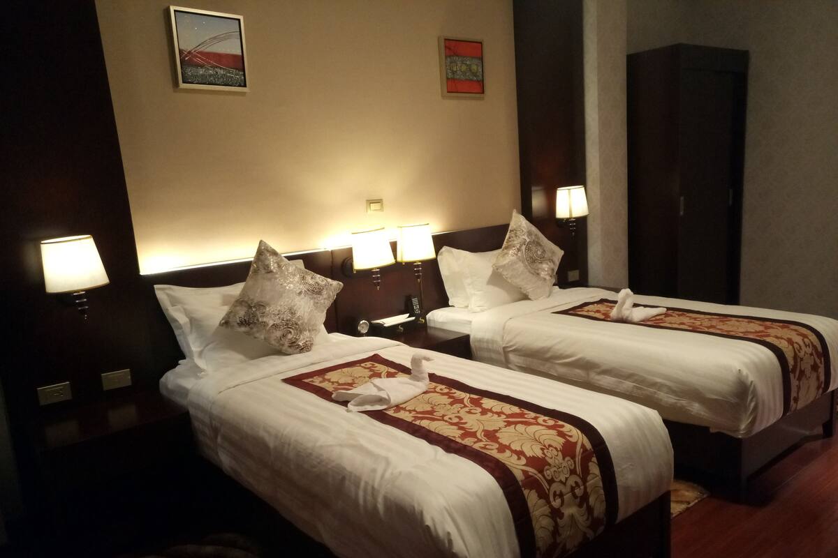 A hotel room with two beds, each with a white bedspread and a red and gold patterned runner. There are bedside lamps, a nightstand with a phone, and a wall-mounted artwork above the beds.
