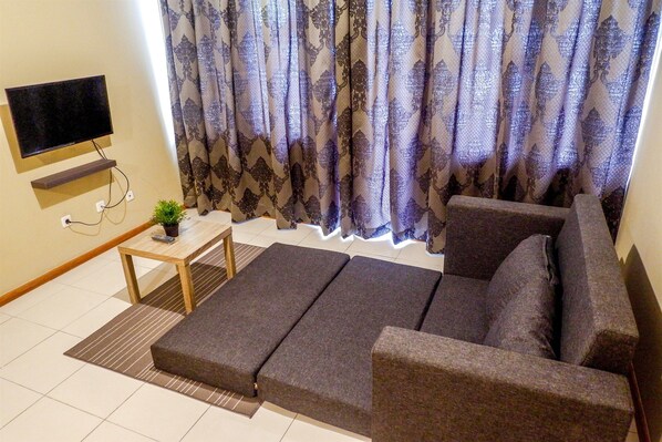 Un salon avec un canapé, un téléviseur fixé au mur, une petite table avec une plante et des rideaux à motifs.