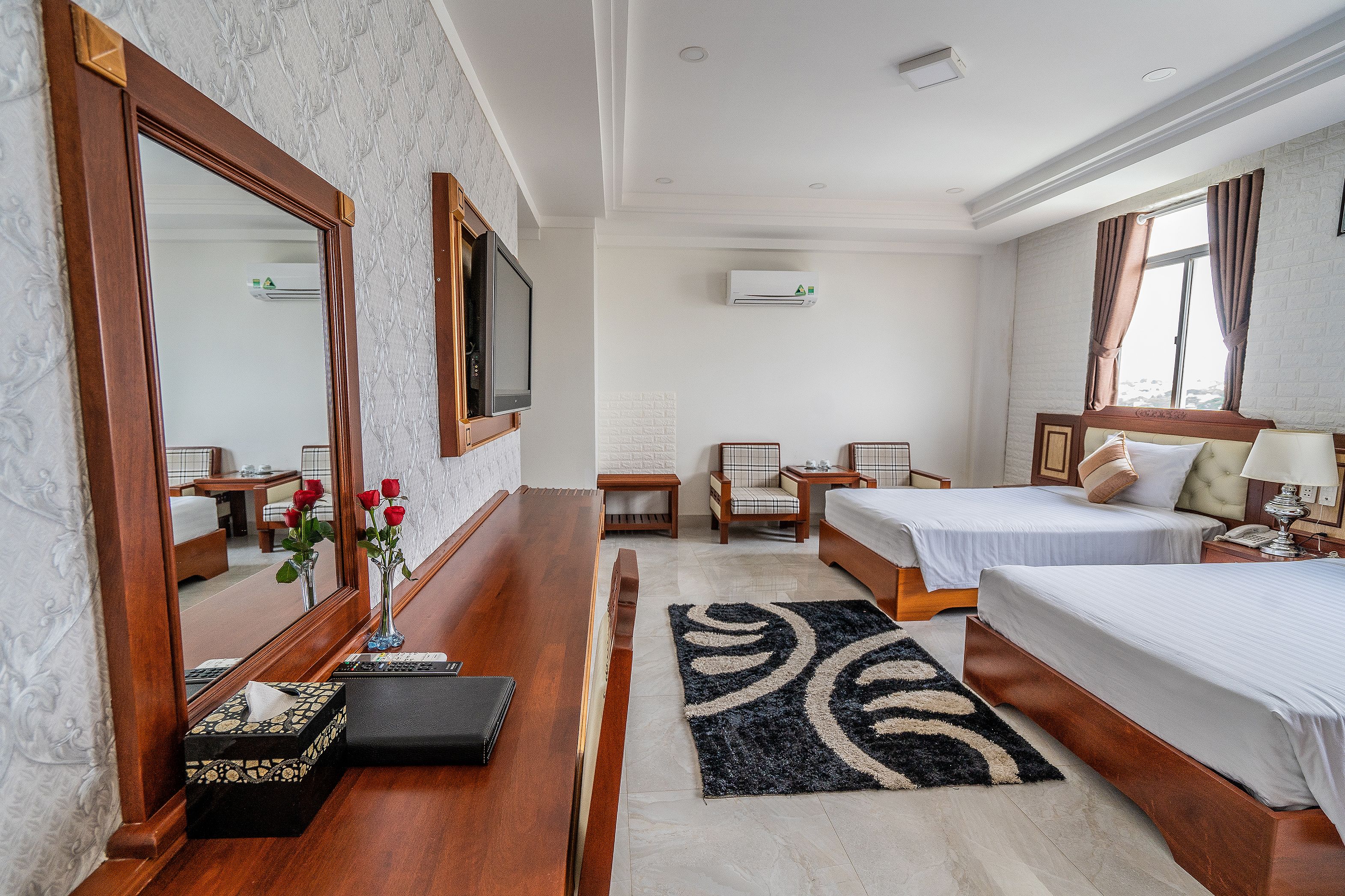 Habitación de hotel con dos camas, televisión, espejo, mesita de noche, lámpara, jarrón con flores, alfombra y aire acondicionado.