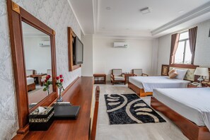 A hotel room with two beds, a TV, a mirror, a bedside table, a nightstand, a lamp, a vase with flowers, a rug, and an air conditioning unit.