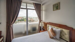 Habitación de hotel con una cama grande, una mesita de noche, una lámpara y vistas a un paisaje urbano por la ventana.