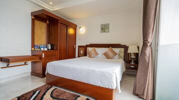 A hotel room with a bed, a wooden wardrobe, a nightstand with a lamp, and a wall-mounted shelf with bottles and a kettle.