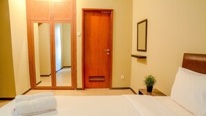 A hotel room with a wooden door, a bed with white linens, a bedside table with a plant, and a mirror on the wall. - 3 Bedrooms Plus Apartment Grand Palace by Travelio (Jakarta)