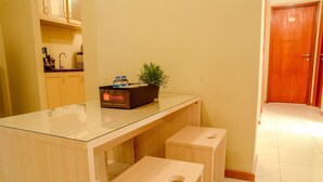 A clean, modern kitchen with a glass-top table, wooden stools, a box labeled 'travello', and a potted plant. - 3 Bedrooms Plus Apartment Grand Palace by Travelio (Jakarta)
