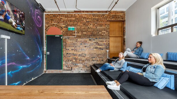 A modern lounge area with brick walls, a large screen, and seating with blue cushions. - Secret Garden Backpackers (Redfern)