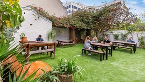 An outdoor patio area with wooden benches and tables, surrounded by lush greenery and potted plants. - Secret Garden Backpackers (Redfern)