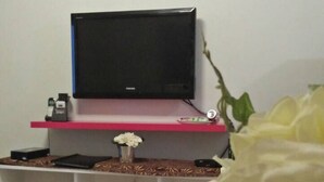 A living room with a flat-screen TV on a pink stand, a white shelf with books and a vase of flowers, and a large white flower in the foreground. - Haven Suite at Jazz (Makati)