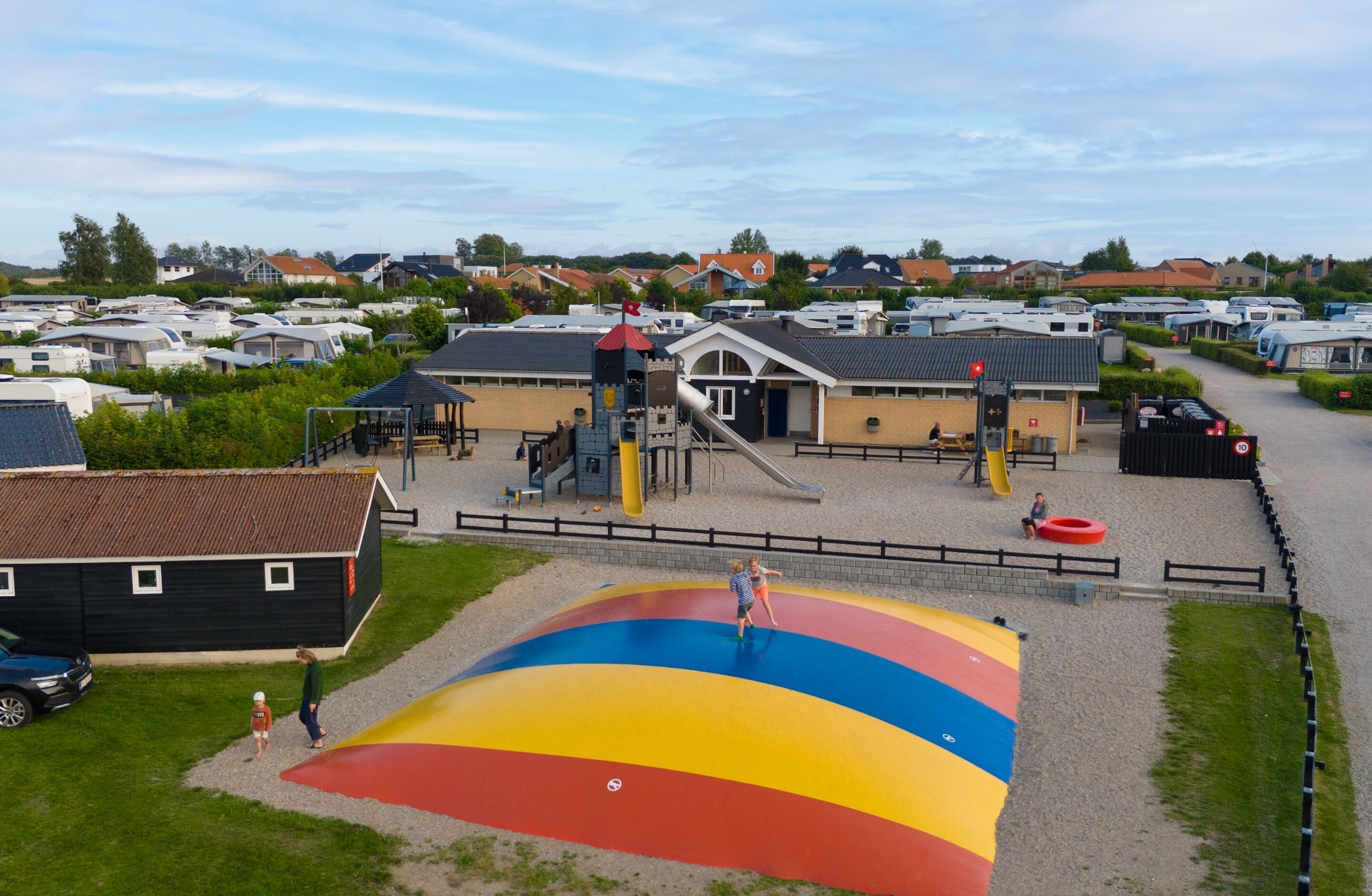 Une aire de jeux avec un toboggan coloré et des structures d’escalade, entourée d’un parking où sont stationnés des camping-cars et d’un petit bâtiment.