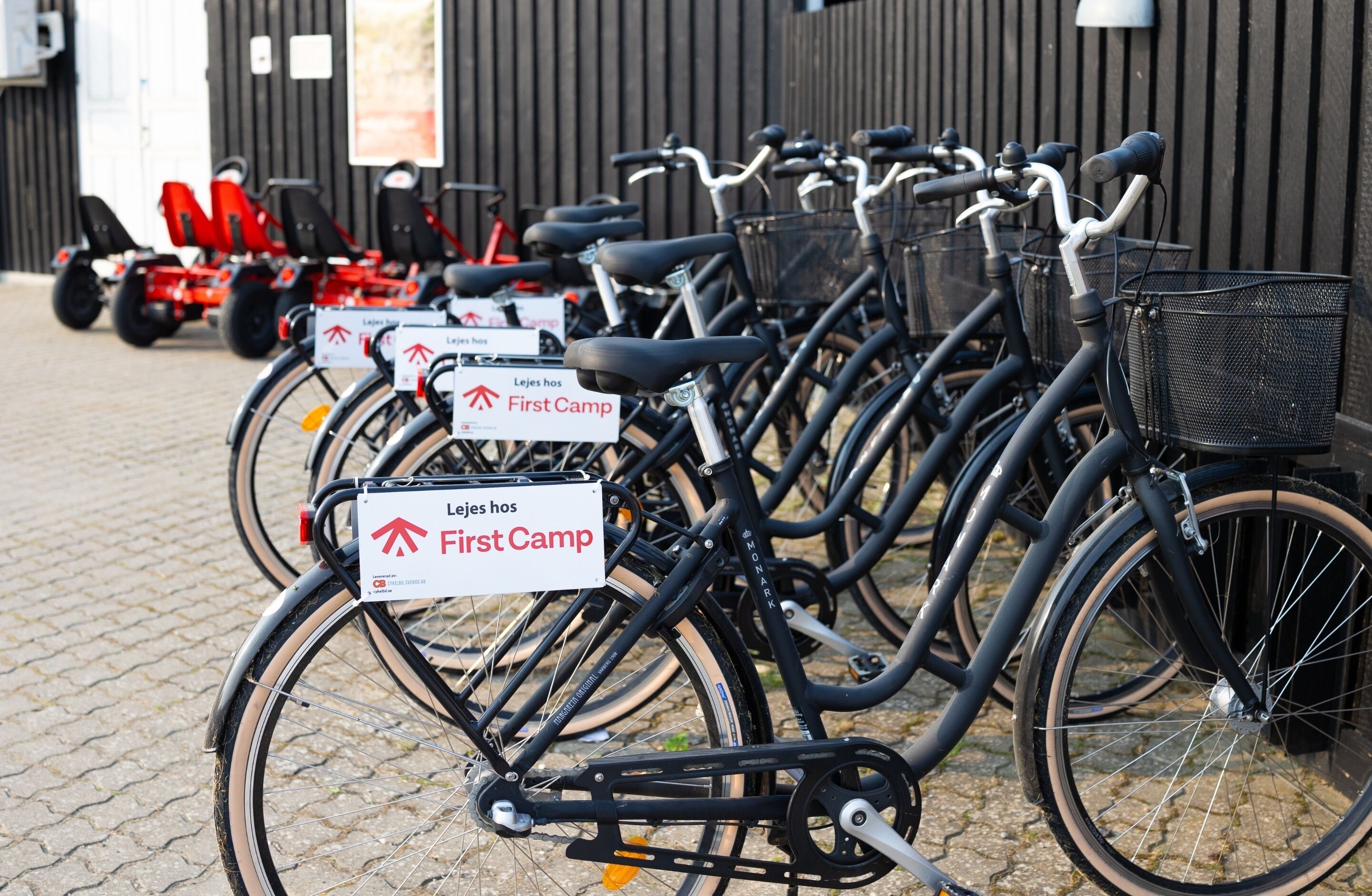 A row of bicycles with "Lejes hos First Camp" signs, parked on a cobblestone surface.