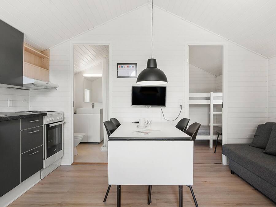 Una cocina compacta con electrodomésticos modernos, una zona de comedor con mesa y sillas, y un sofá gris en una habitación luminosa con paredes blancas.