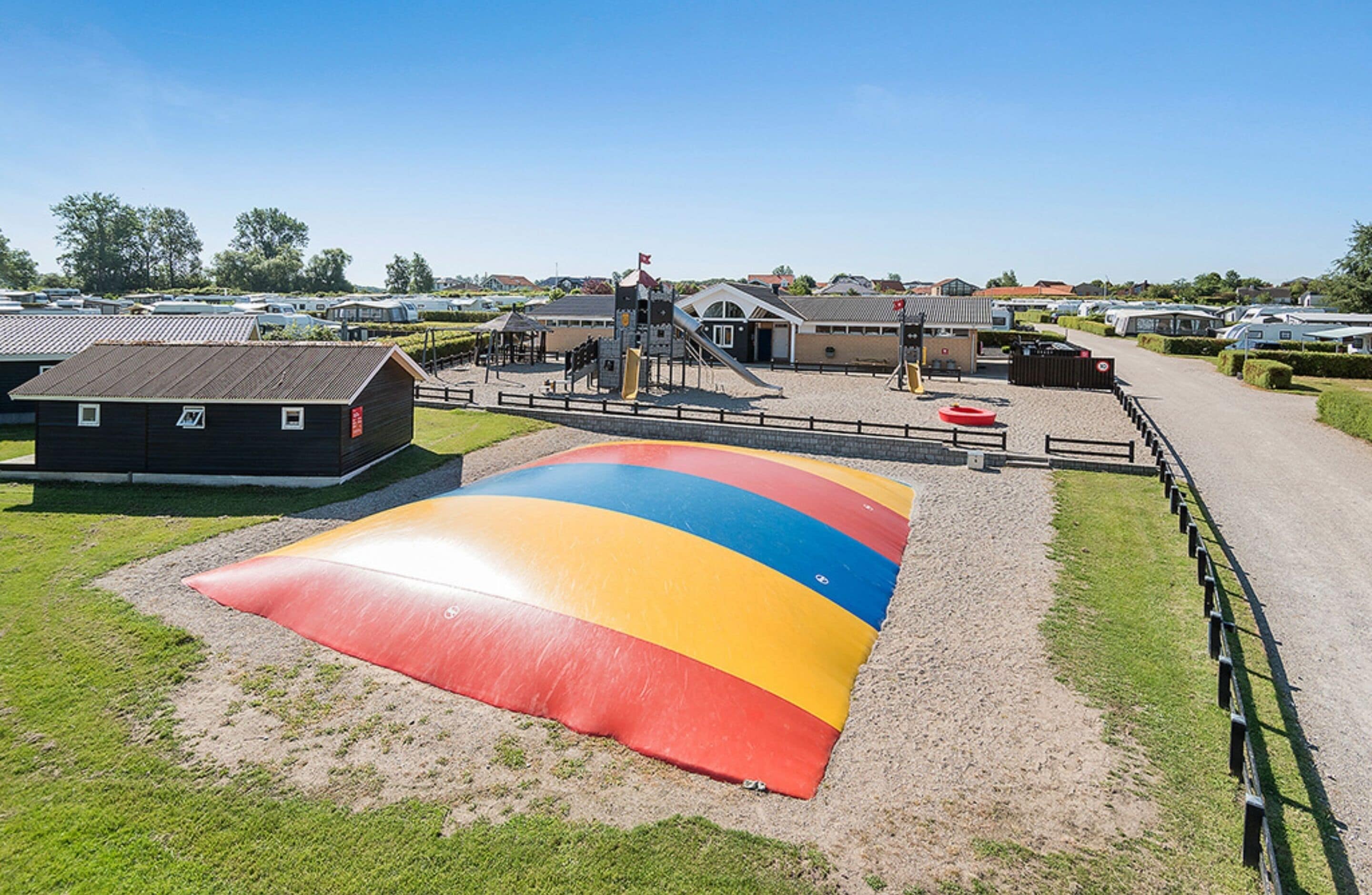 A large inflatable slide in a park area with a playground, a small building, and a parking lot with RVs in the background.