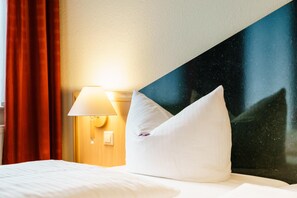 Une chambre d’hôtel avec un lit, des oreillers blancs, une table de chevet avec une lampe et une grande fenêtre donnant sur le ciel.