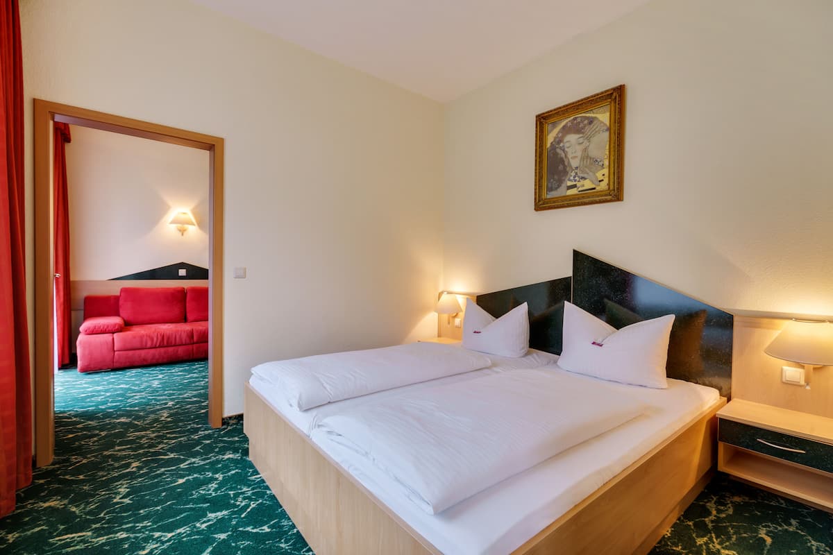 A hotel room with a large bed, two bedside lamps, a nightstand, and a red sofa in the adjacent room.