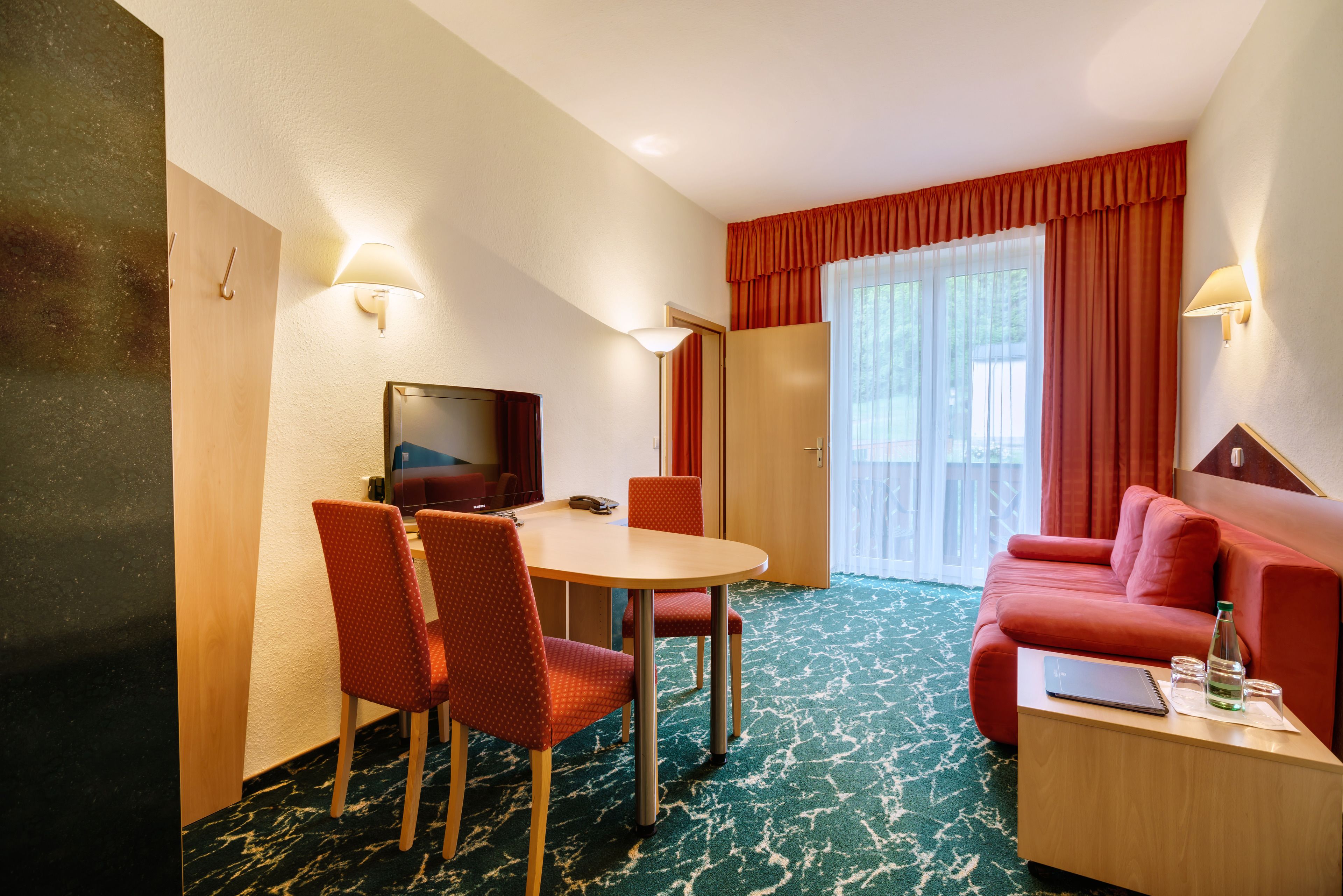 Habitación de hotel con una mesa de comedor, sillas, televisor, sofá y ventana con cortinas.
