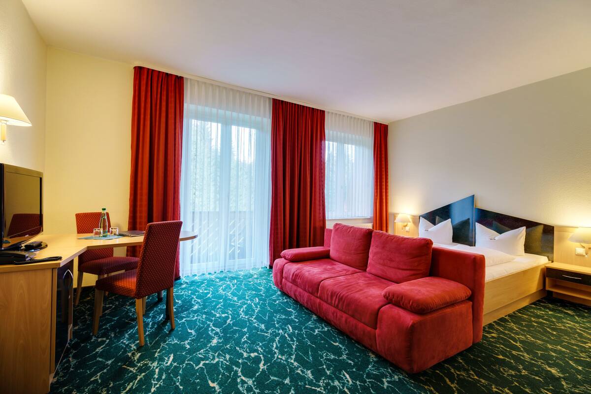 A hotel room with a bed, a red sofa, a desk with a chair, and a TV.