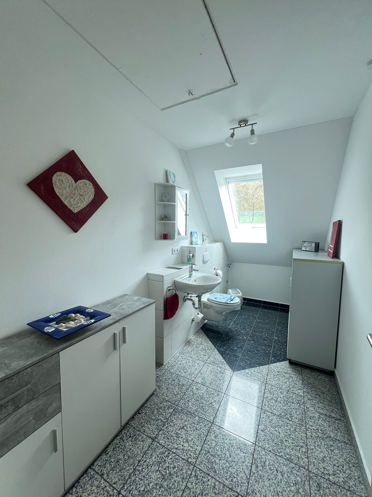 A small, modern bathroom with a white sink, a toilet, a cabinet, and a window.