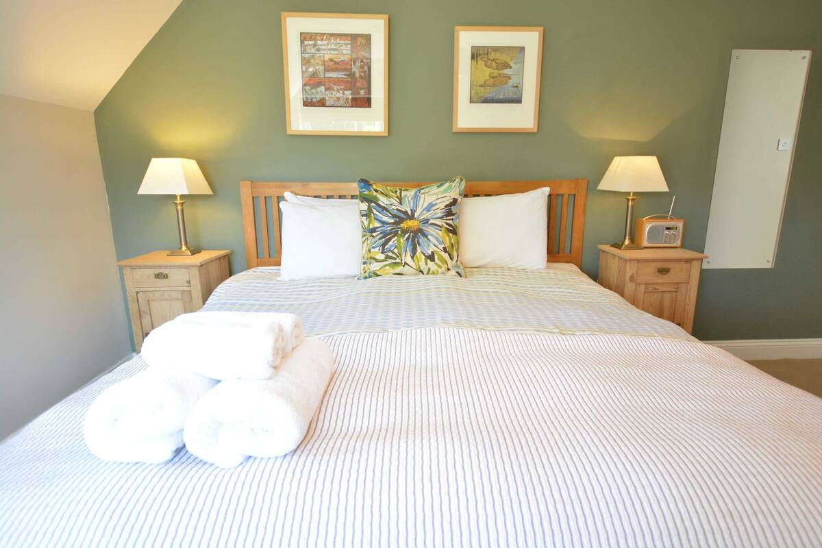 A neatly made bed with striped bedding, two pillows, a floral pillow, and a set of white towels. There are two bedside tables with lamps and a radio. The room has a green wall with two framed pictures above the bed.