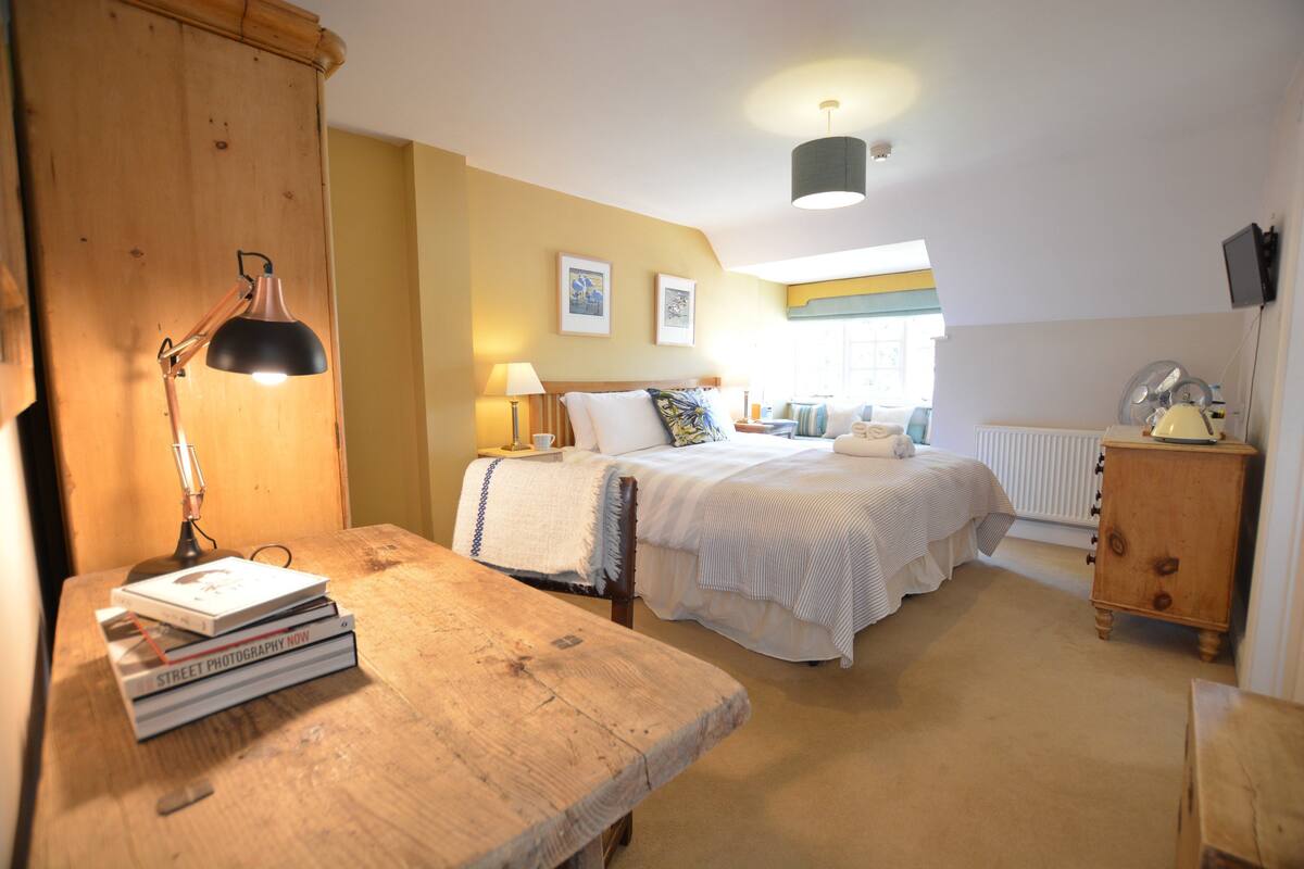 A bedroom with a wooden desk, books, a lamp, a bed with a white bedspread, and a wooden cabinet.