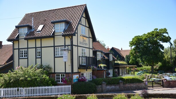 A traditional Tudor-style building with a sign reading "The Dolphin." It features a white picket fence, a grassy area with yellow flowers, and a paved walkway. - The Dolphin Inn (Leiston)