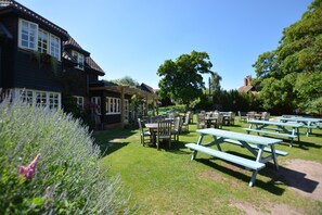 A garden with picnic tables and chairs, a building with windows and a balcony, and a large tree in the background.
