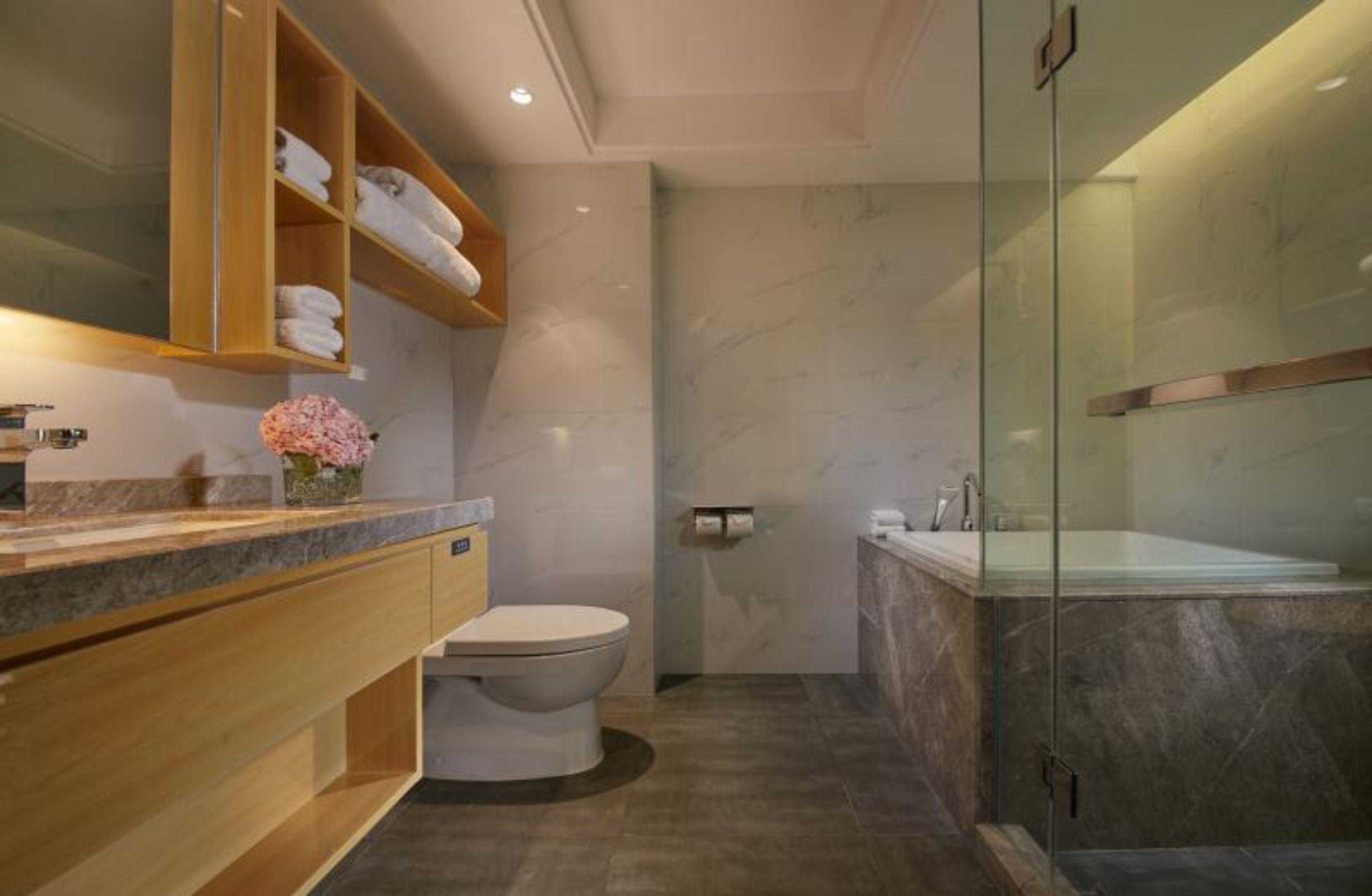 Un baño moderno con suelo de mármol, cabina de ducha de vidrio, tocador de madera con lavabo e inodoro.