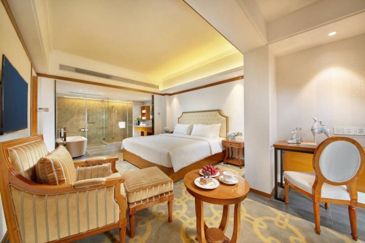 Habitación de hotel con una cama grande, una zona de estar con sofá y sillón, un escritorio con silla, y una mesita con una fuente de fruta y una taza.