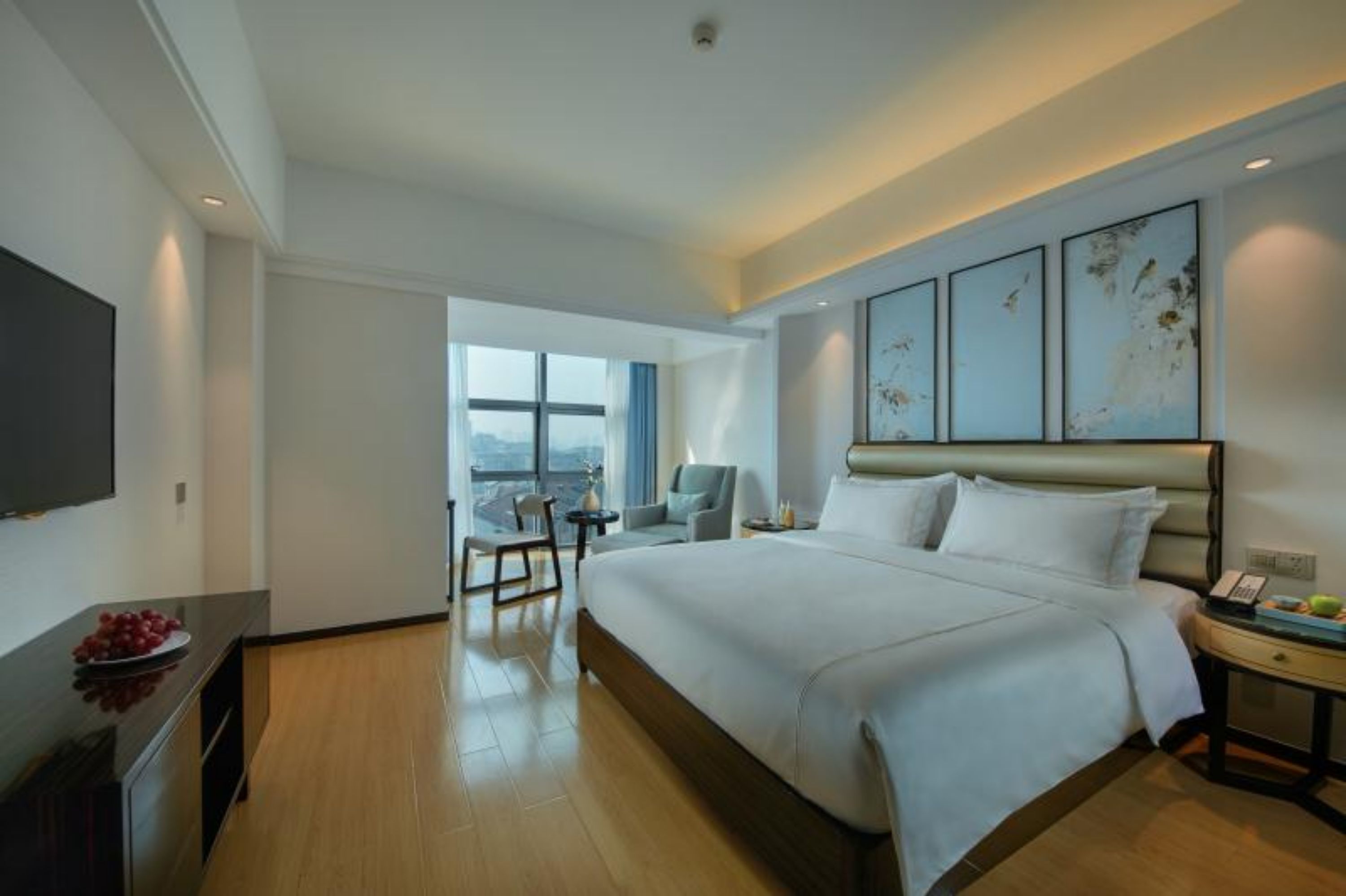 A modern hotel room with a large bed, a flat-screen TV, a dining table, and a view of the city.