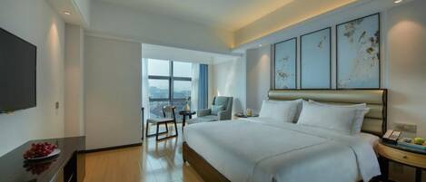 A modern hotel room with a large bed, a flat-screen TV, a dining table, and a view of the city.
