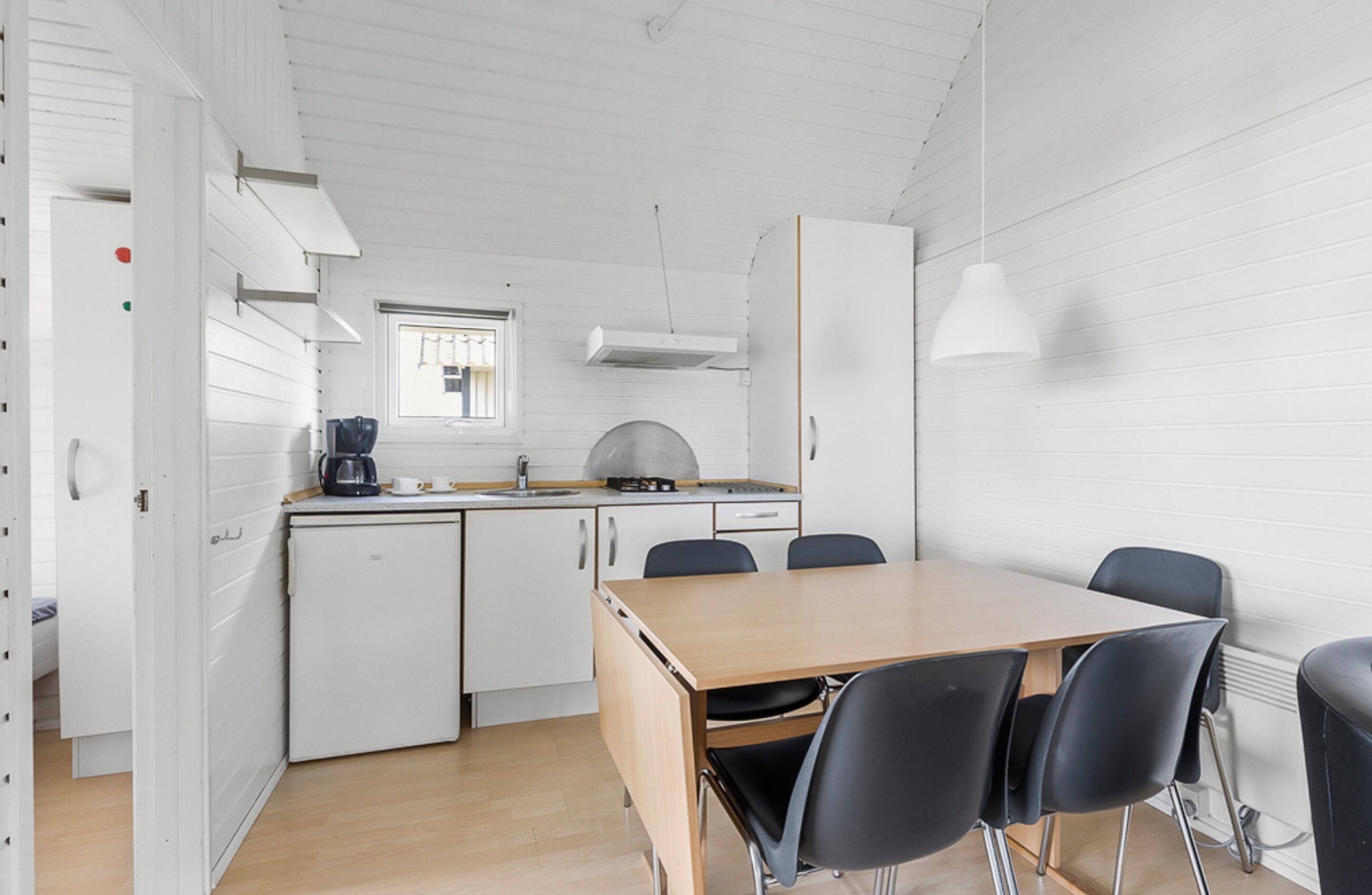 Una cocina compacta con comedor, muebles blancos, un refrigerador pequeño y una ventana con rejas.