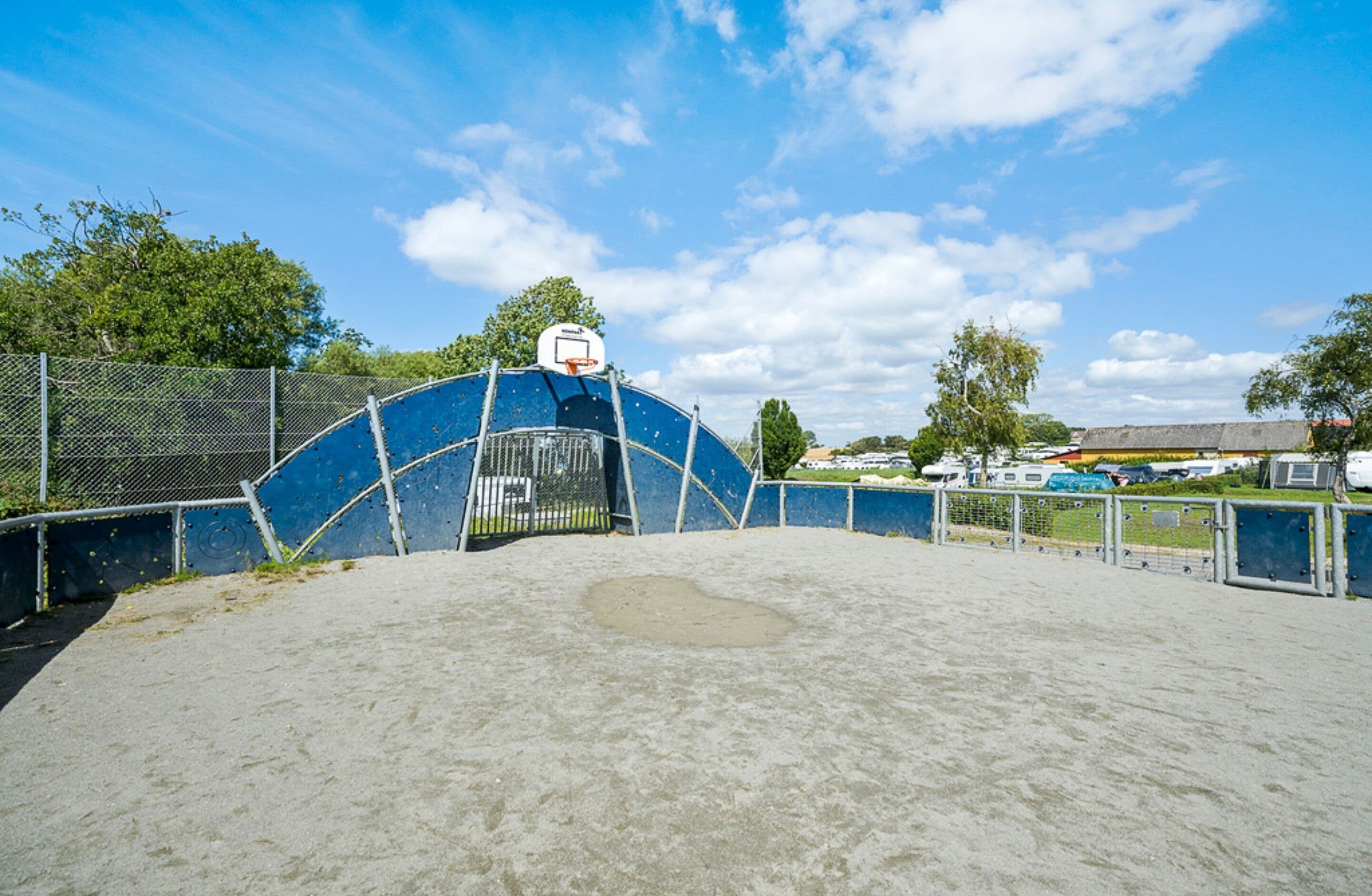 Uma quadra de basquete ao ar livre com cercas azuis, um aro de basquete e superfície de areia.