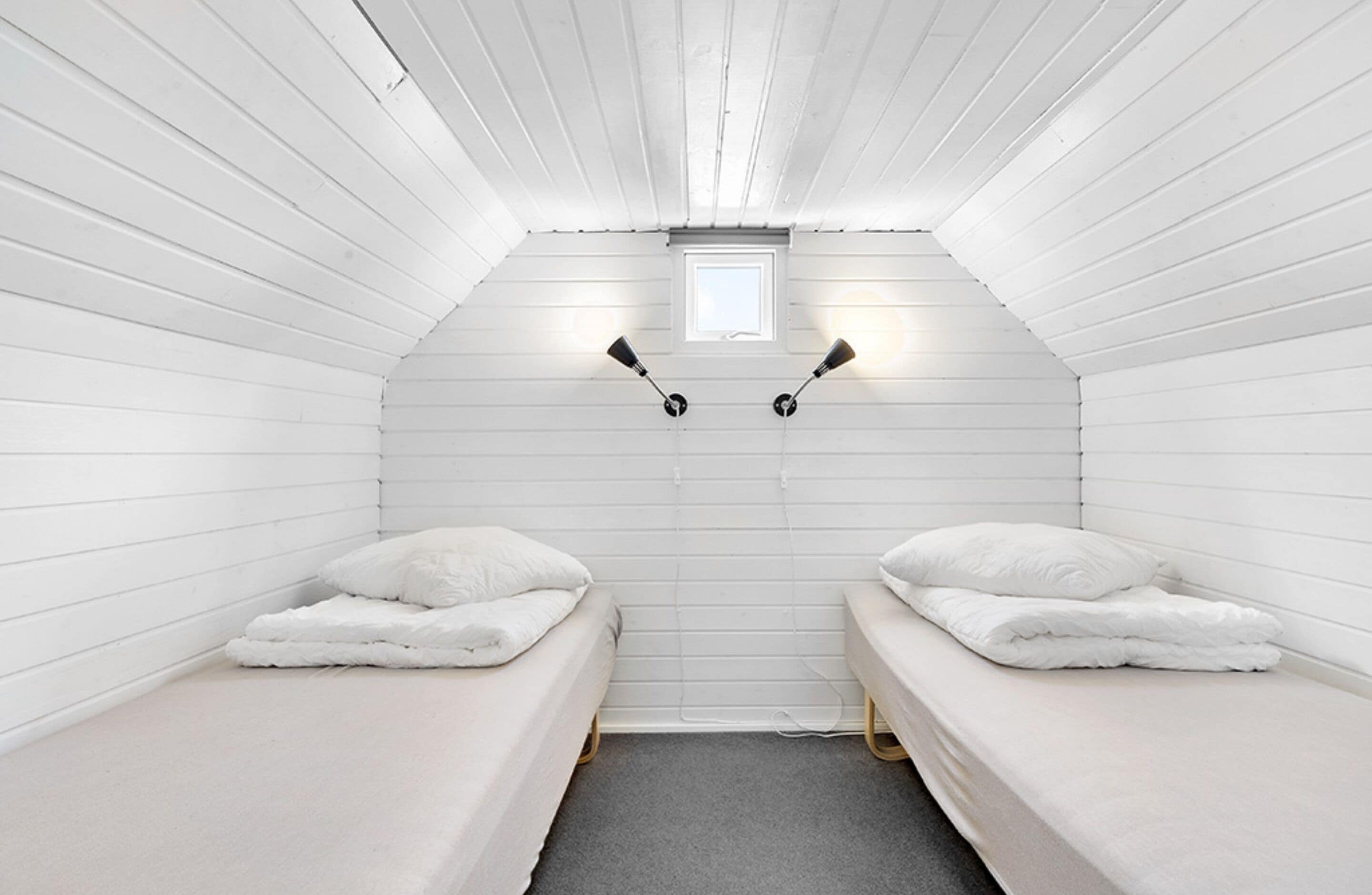 A small, white-paneled room with two single beds, a small window, and two wall-mounted lamps.