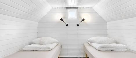 A small, white-paneled room with two single beds, a small window, and two wall-mounted lamps.