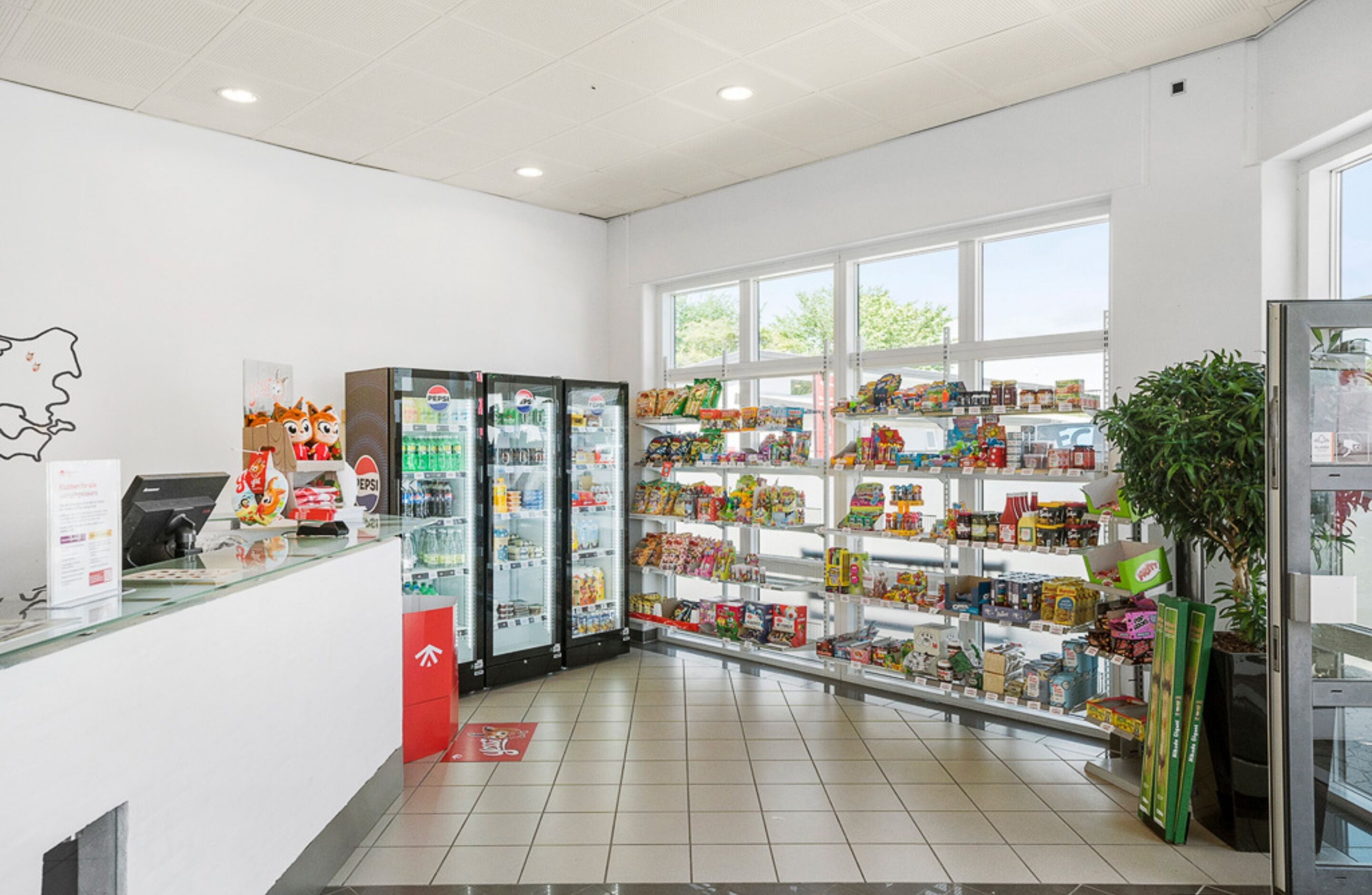 L’intérieur d’un magasin, avec un comptoir de caisse, des distributeurs de boissons réfrigérées, des étagères remplies d’en-cas et une grande fenêtre laissant entrer la lumière naturelle.