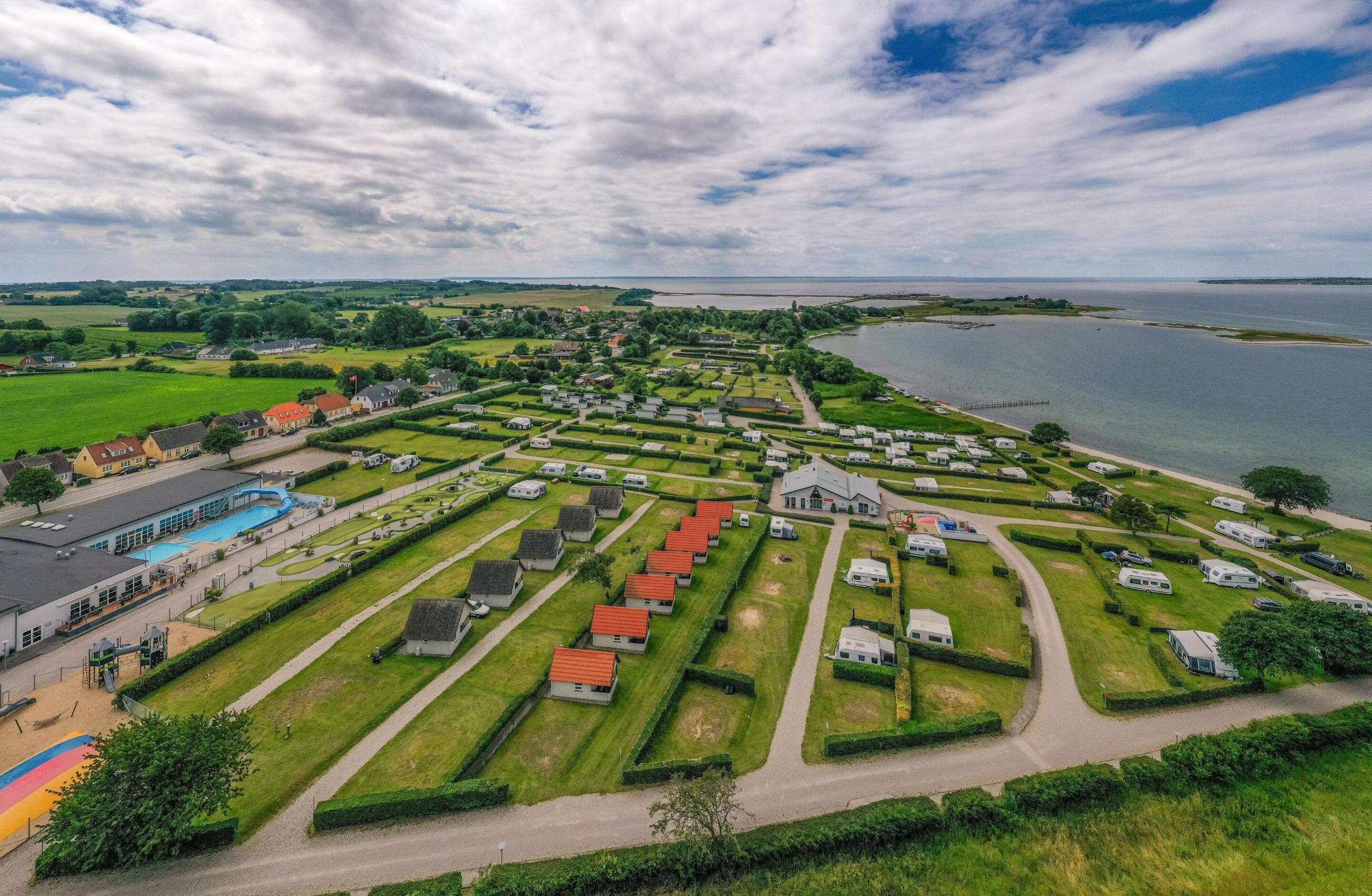 Vista aérea de un camping junto al lago con vehículos recreativos y cabañas ordenados, una piscina y un muelle que se adentra en el agua.