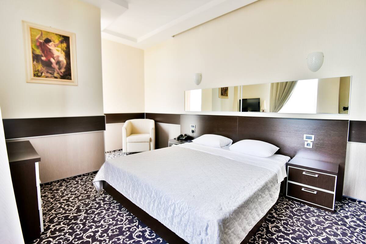 A hotel room with a large bed, a bedside table, a chair, a desk, a mirror, and a painting on the wall.