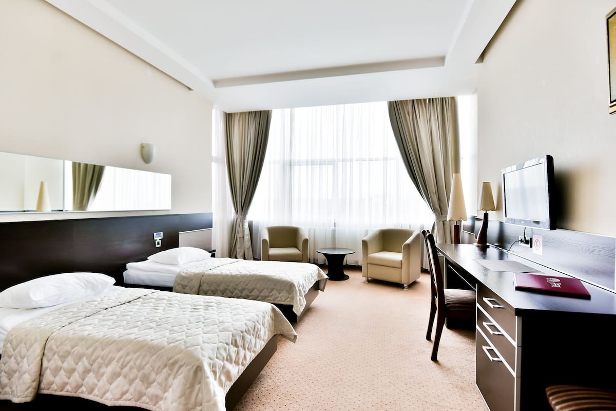 A hotel room with two beds, a desk, a chair, a TV, and a large window with curtains.