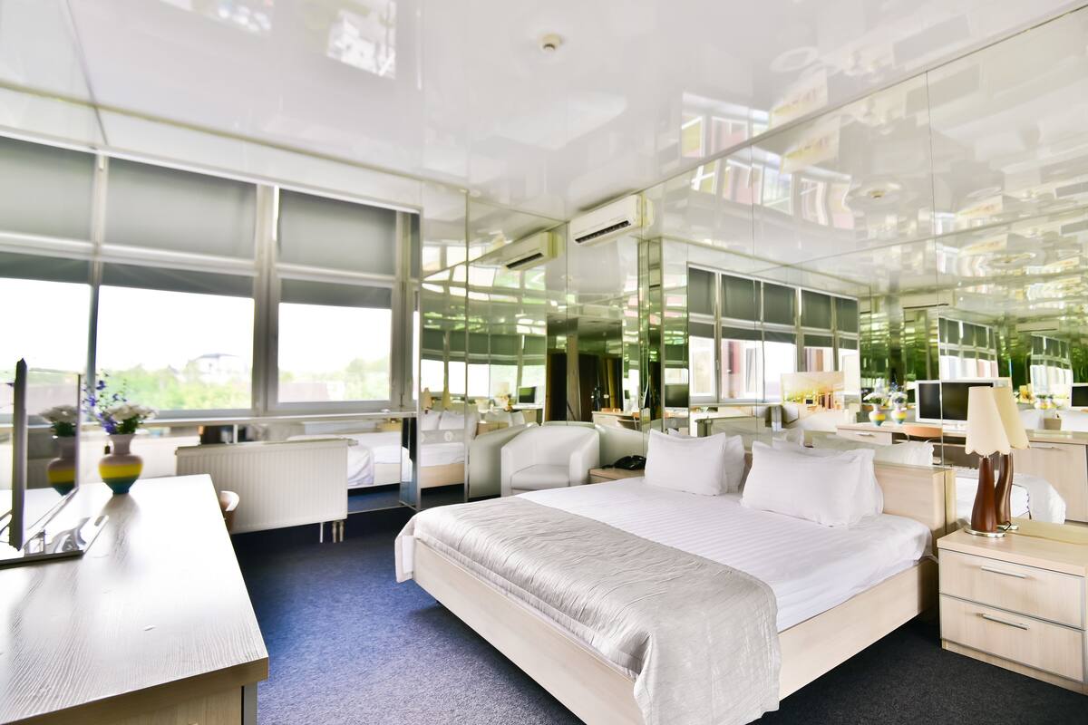 A modern hotel room with a large bed, a desk, and a chair. There are mirrors on the walls and a window providing a view of the outdoors.