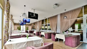 Un restaurant doté de nombreuses tables et chaises, de grandes fenêtres avec des rideaux et d’un écran de télévision.
