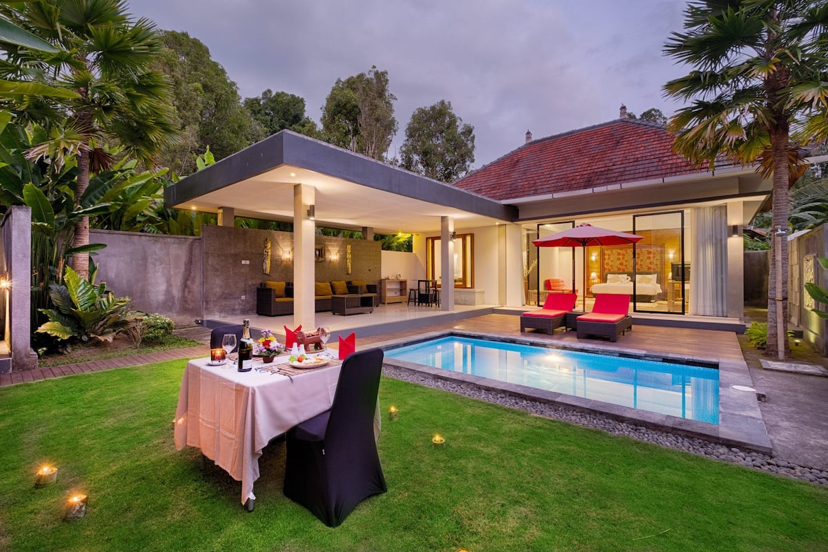 A modern outdoor dining area with a pool, covered seating, and a patio with a red umbrella.