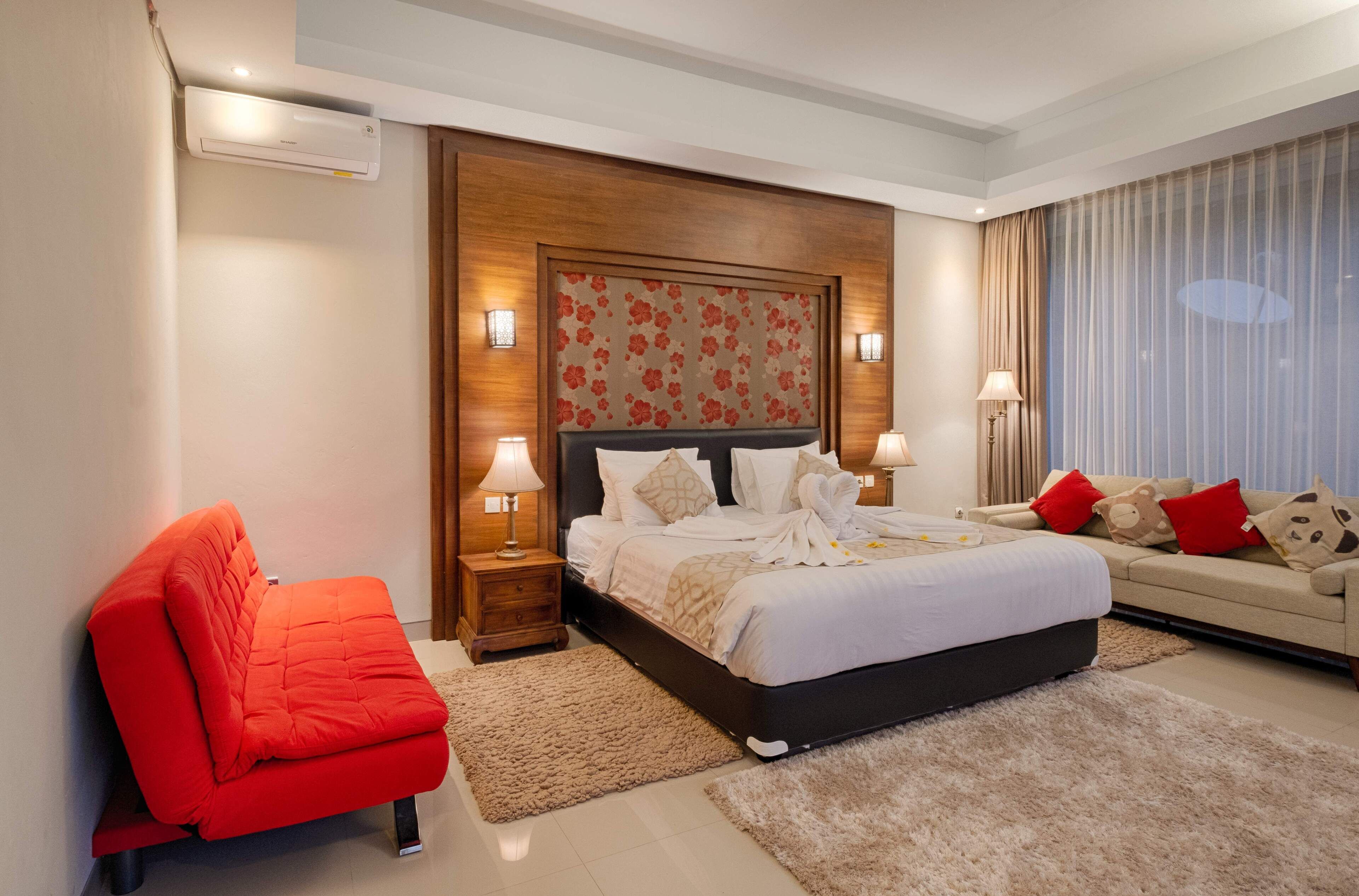 Une chambre d’hôtel avec un lit, un fauteuil rouge, un canapé et une grande fenêtre avec des rideaux.