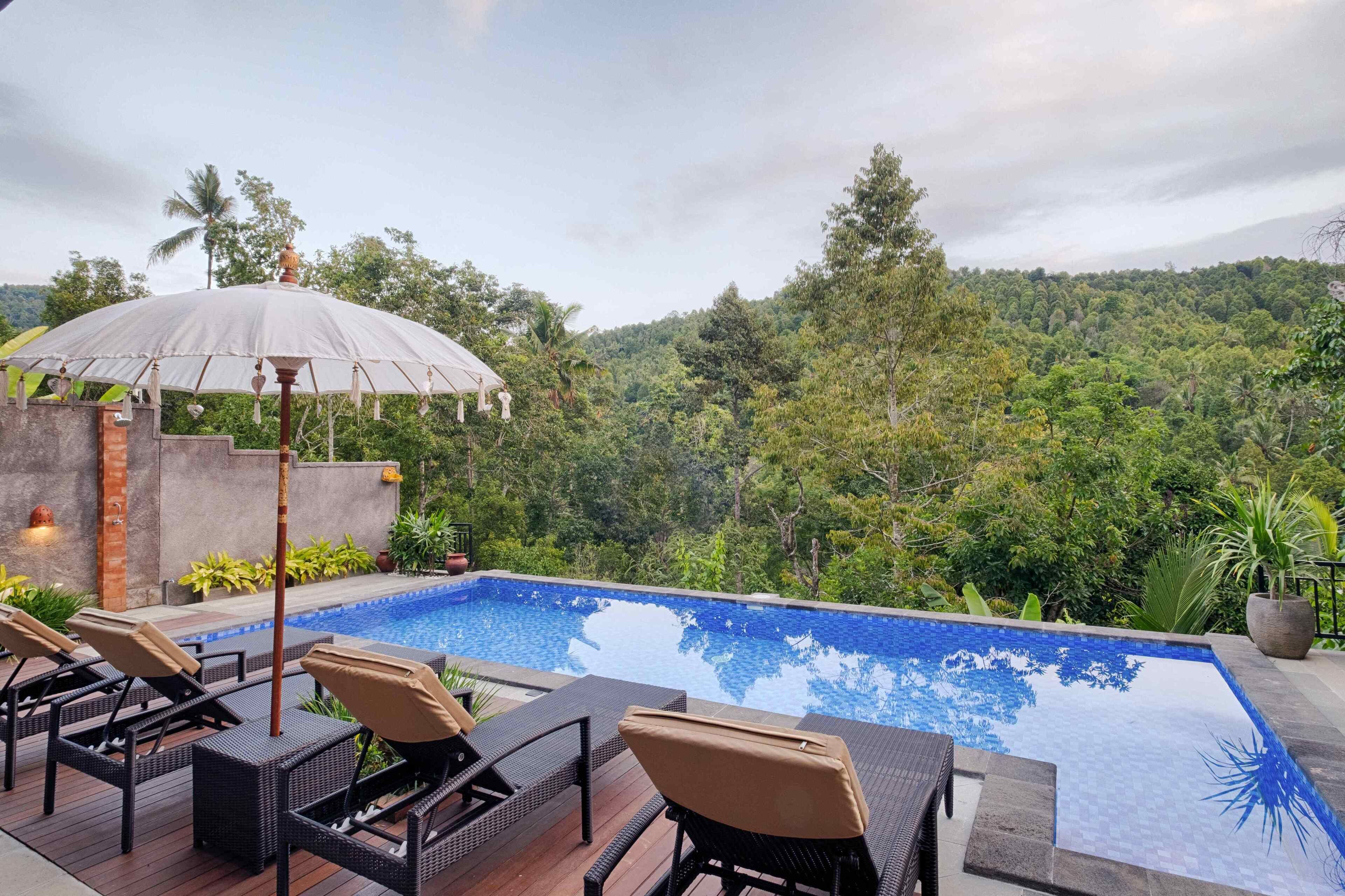 A pool with lounge chairs and an umbrella, surrounded by lush greenery and a mountainous backdrop.