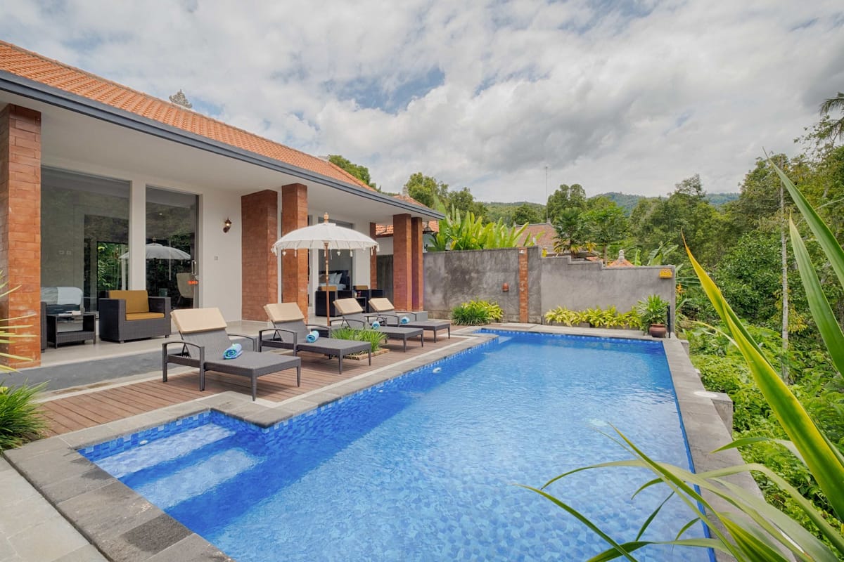 A swimming pool with lounge chairs and umbrellas, surrounded by greenery and a brick building.